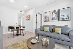 Ein modernes Wohnzimmer mit einem grauen Sofa, einem Esstisch und Stühlen. An den Wänden hängen gerahmte Bilder, auf dem Couchtisch befinden sich eine Schale und eine Vase, und ein Teppich liegt auf dem Boden.