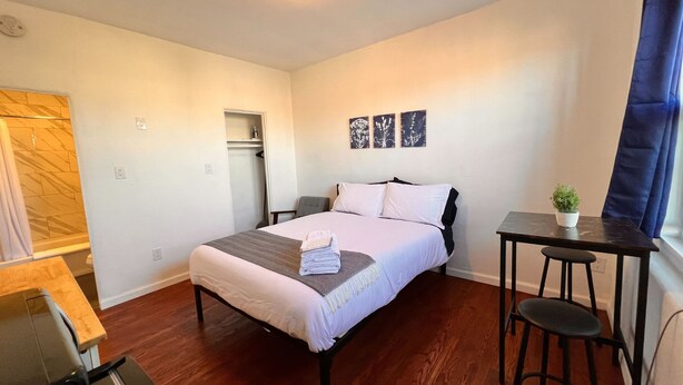 Quarto com cama, escrivaninha, cadeira, pequena mesa com planta e área de chuveiro.