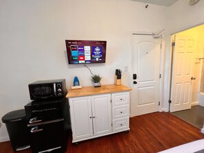 Ein Zimmer mit einem an der Wand montierten Flachbildfernseher, der verschiedene Streaming-Dienste anzeigt, einem schwarzen Kühlschrank, einem weißen Schrank mit Schubladen und einer Holz Arbeitsplatte.