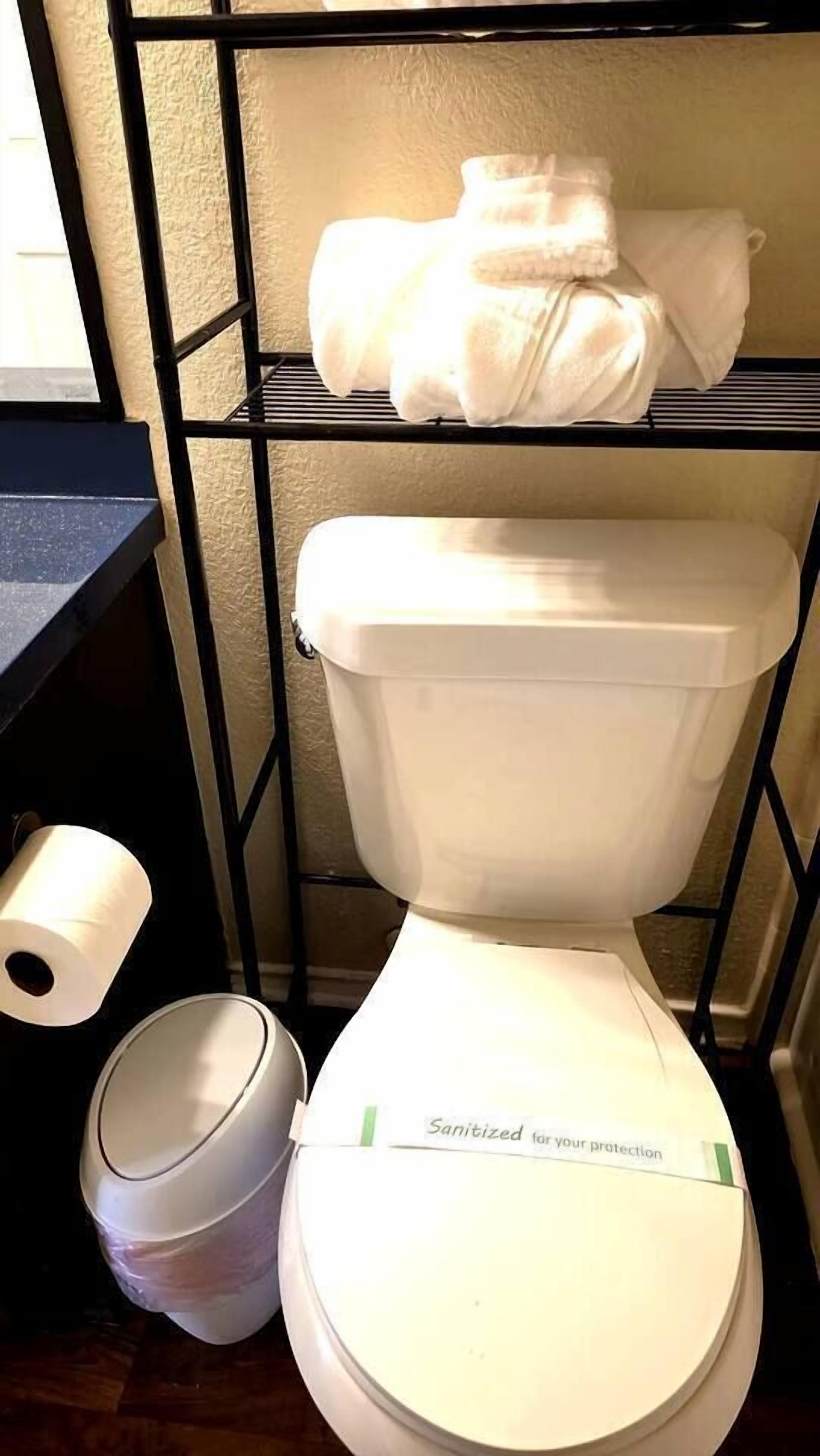 A toilet with a lid that has a 'Sanitized for your protection' label, a toilet paper roll, and a trash bin.