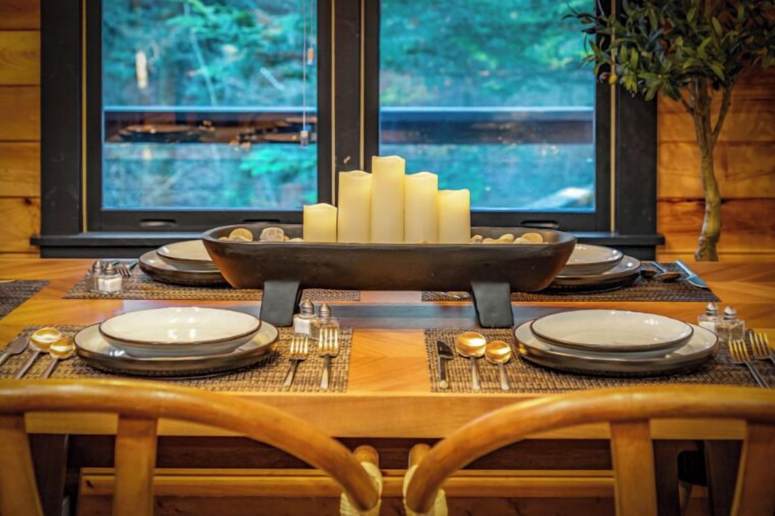 A dining table set with plates, cutlery, and candles, with a window view of a forest.