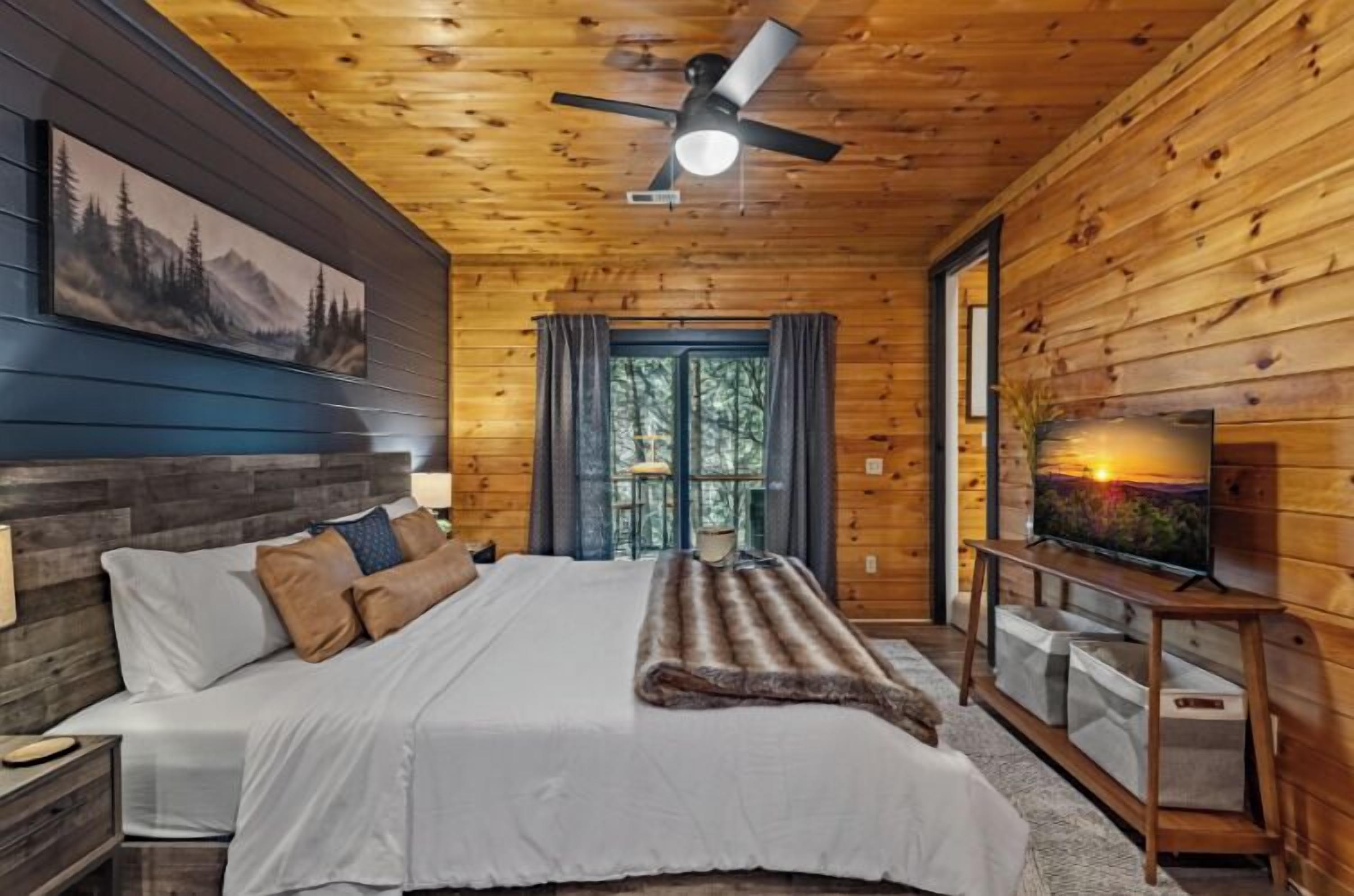 A wooden cabin bedroom with a bed, a ceiling fan, a TV, and a window.