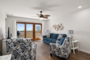 A living room with a blue sofa, patterned armchairs, a ceiling fan, and a view of the beach through glass doors. - Gulf-view 1BR condo at La Mirage with pool, hot tub, and beach access. (Port Aransas)