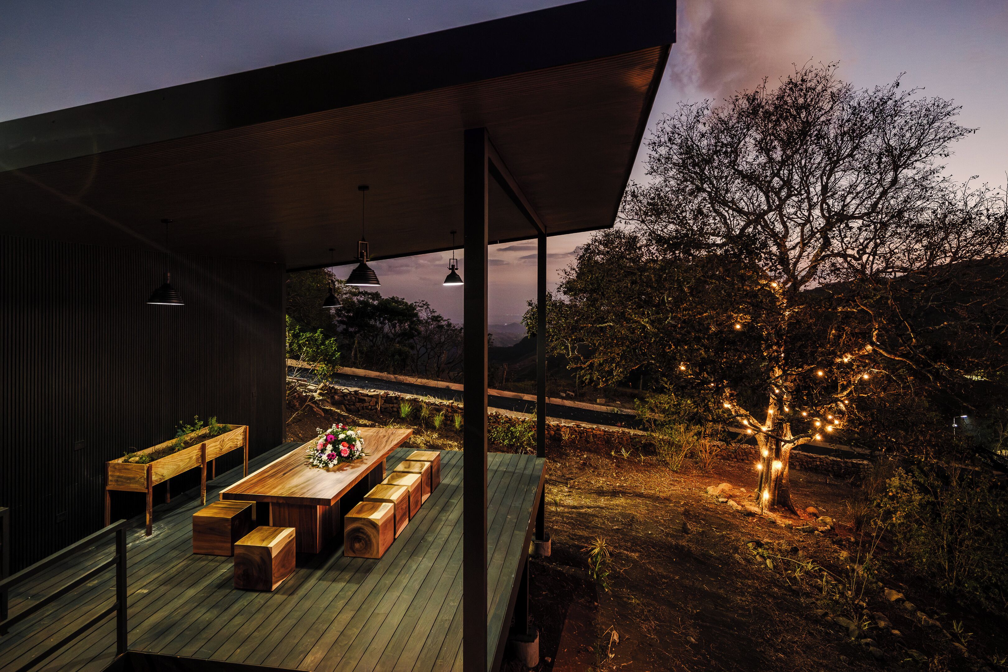 Una terrazza esterna con tavolo e panche in legno, affacciata su un giardino curato con un albero illuminato da luci decorative.