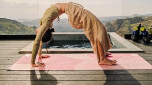 Iemand doet een yoga-houding op een roze mat, buiten, met een zwembad en bergen op de achtergrond.
