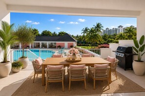 Zona de comedor junto a la piscina, con mesa de madera, sillas de mimbre y vista a un hotel y palmeras.