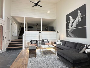 A modern living room with a sofa, coffee table, and chairs, a staircase leading to an upper level, and a large abstract artwork on the wall. - 2BR/2BA Whitefish Lake Overlook at Suncrest (Whitefish)