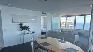 Ett modernt hotellrum med ett stort ovalt bord, grå stoppade sittmöbler, ett skrivbord i glas och utsikt över havet genom stora fönster.