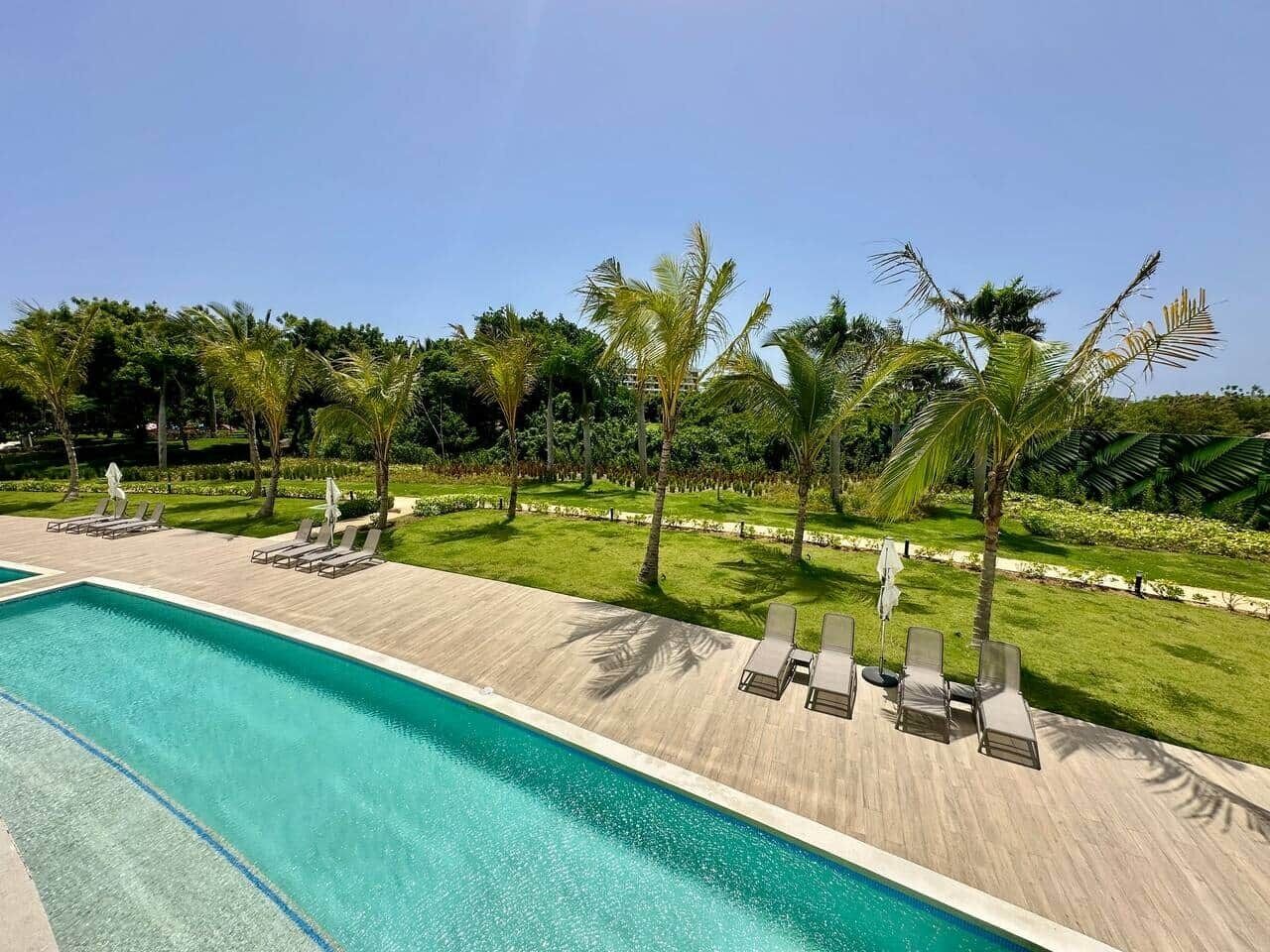 A swimming pool with lounge chairs and umbrellas, surrounded by palm trees and a well-maintained lawn.