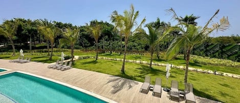 A swimming pool with lounge chairs and umbrellas, surrounded by palm trees and a well-maintained lawn.