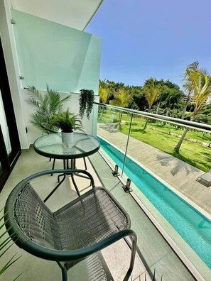 A balcony with a glass table and chairs, overlooking a swimming pool and a well-maintained lawn. - Cana Life Luxury | Cana Rock Universe Pool View (Punta Cana)