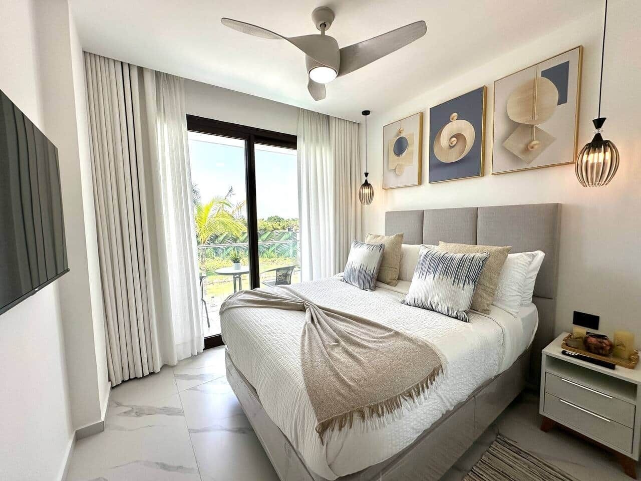 Een moderne slaapkamer met een groot bed, een plafondventilator, een aan de muur bevestigde televisie en kunst boven het bed.