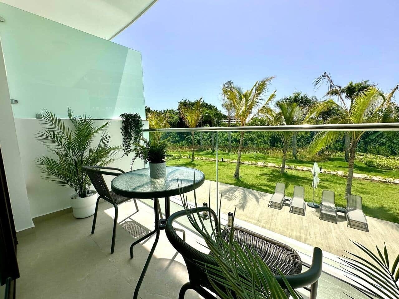 Un balcon agrémenté d’une table et de chaises en verre, entouré de plantes en pot, et offrant une vue sur une pelouse bien entretenue et des palmiers.