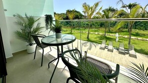 Uma varanda com mesa e cadeiras de vidro, rodeada por plantas em vasos e vista para um gramado bem cuidado e palmeiras.