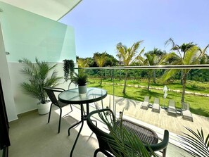 A balcony with a glass table and chairs, surrounded by potted plants and a view of a well-maintained lawn and palm trees. - Cana Life Luxury | Cana Rock Universe Pool View (Punta Cana)