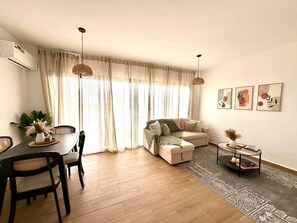 Ein Wohnzimmer mit Esstisch, Sofa und Couchtisch, ausgestattet mit einem großen Fenster und Vorhängen.