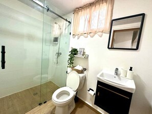 A bathroom with a glass shower enclosure, a toilet, a sink with a mirror above it, and a shelf with toiletries. - Cana Life Breeze | Blue Coral Residences C2 (Punta Cana)