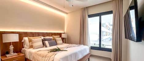 A modern hotel room with a large bed, a ceiling fan, a TV mounted on the wall, and a window with curtains.