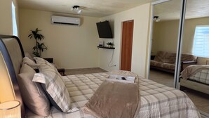 Ein Schlafzimmer mit einem Bett, einem an der Wand befestigten Fernseher, einer Pflanze und einer Schiebetür zu einem weiteren Raum.