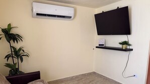 Ein Wohnzimmer mit flachbildfernseher an der Wand, einer Topfpflanze, einer kleinen Ablage mit einem Schild und einem beigefarbenen Sofa.