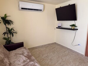 A living room with a flat-screen TV mounted on the wall, a potted plant, a small shelf with a sign, and a beige couch. - Description (Fajardo)