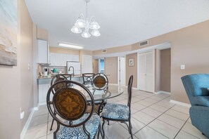 A dining area with a glass-top table and chairs, a kitchen in the background, and a hallway leading to a door. - Gulf Shores Plantation Palms 6603 | Pool + Hot Tub (Gulf Shores)