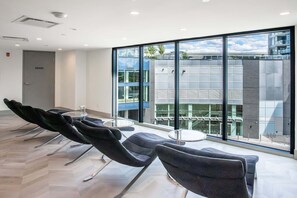 A modern waiting area with a row of chairs, a small table, and large windows offering a view of a building with a glass facade. - The Aqua Collection - Lakeview Terrace - 902 (Kelowna)