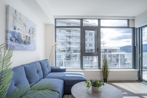 A modern living room with a blue sofa, a round coffee table, and a large window offering a view of a building complex. - The Aqua Collection - Lakeview Terrace - 902 (Kelowna)