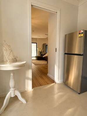 Een keuken met een roestvrijstalen koelkast, een witte bijzettafel en een doorgang naar een woonkamer met een bank en een vloerkleed.