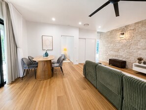 Een moderne woonkamer met een houten vloer, een ronde eettafel met stoelen, een groene bank, een accentmuur van steen en een witte console tafel.