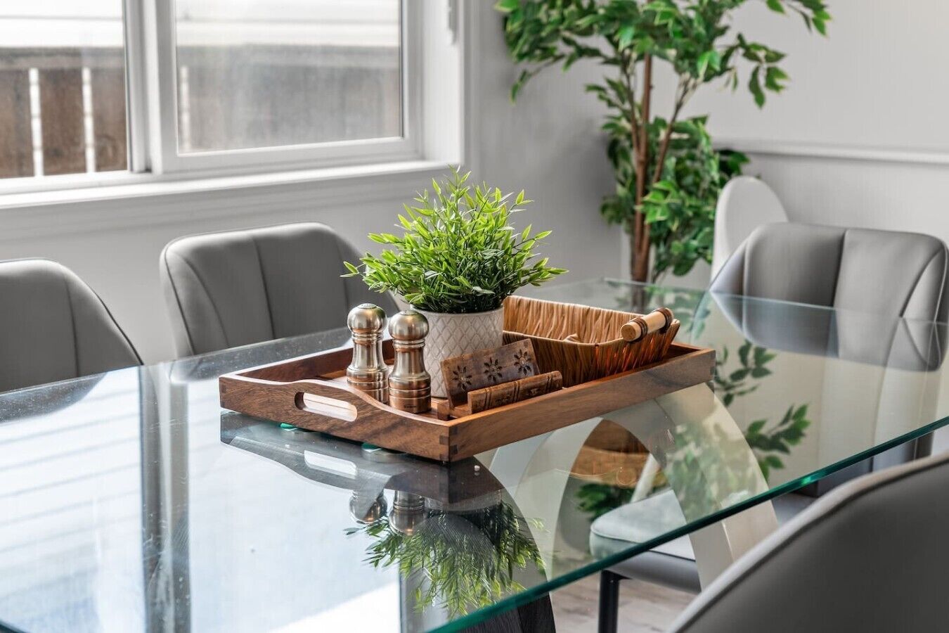 Una mesa de comedor moderna con superficie de vidrio, rodeada por sillas grises. En la mesa, una bandeja de madera con una planta en maceta y saleros y pimenteros.