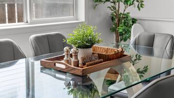 Una mesa de comedor moderna con superficie de vidrio, rodeada por sillas grises. En la mesa, una bandeja de madera con una planta en maceta y saleros y pimenteros.