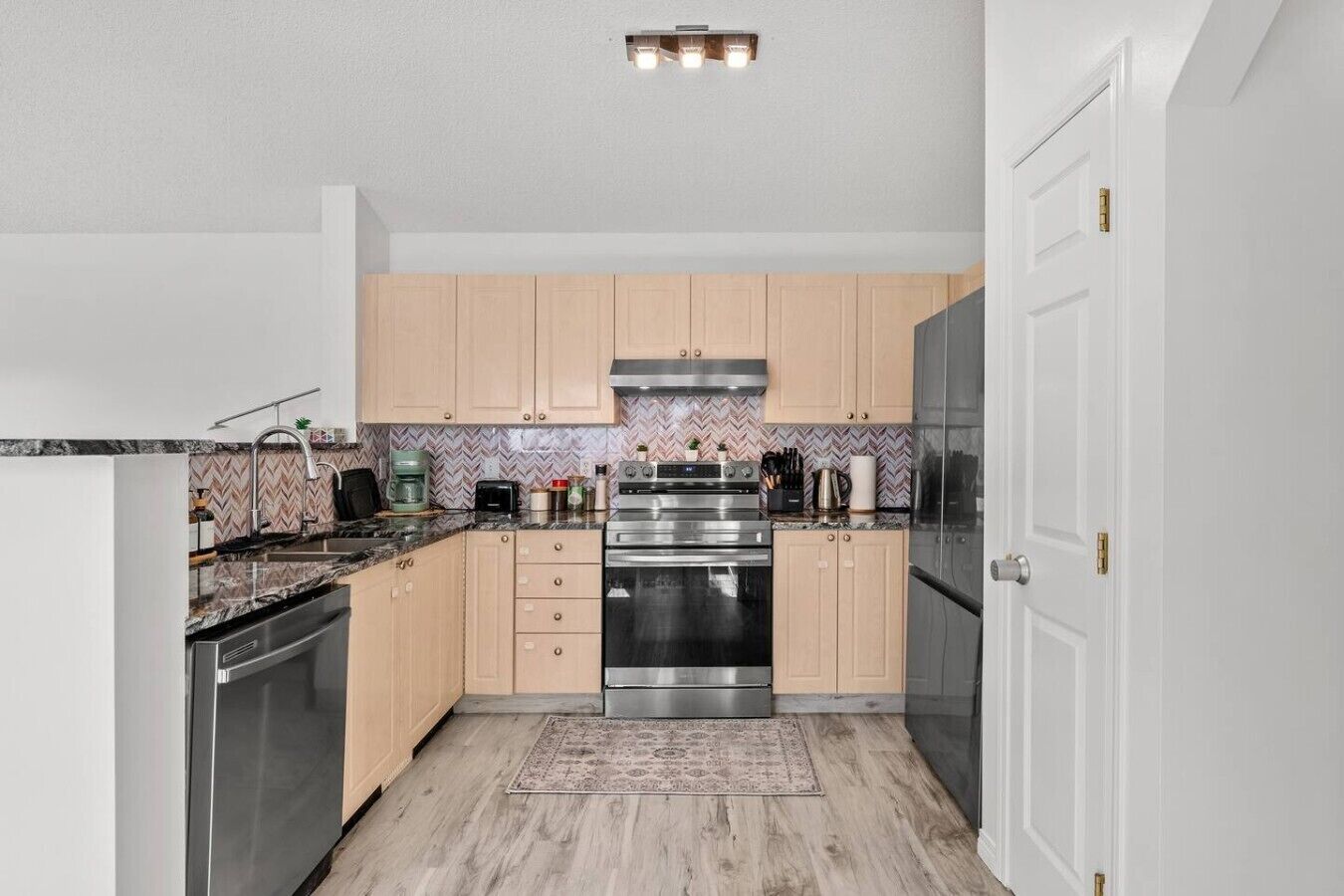 A modern kitchen with light wood cabinets, stainless steel appliances, and a herringbone patterned backsplash.