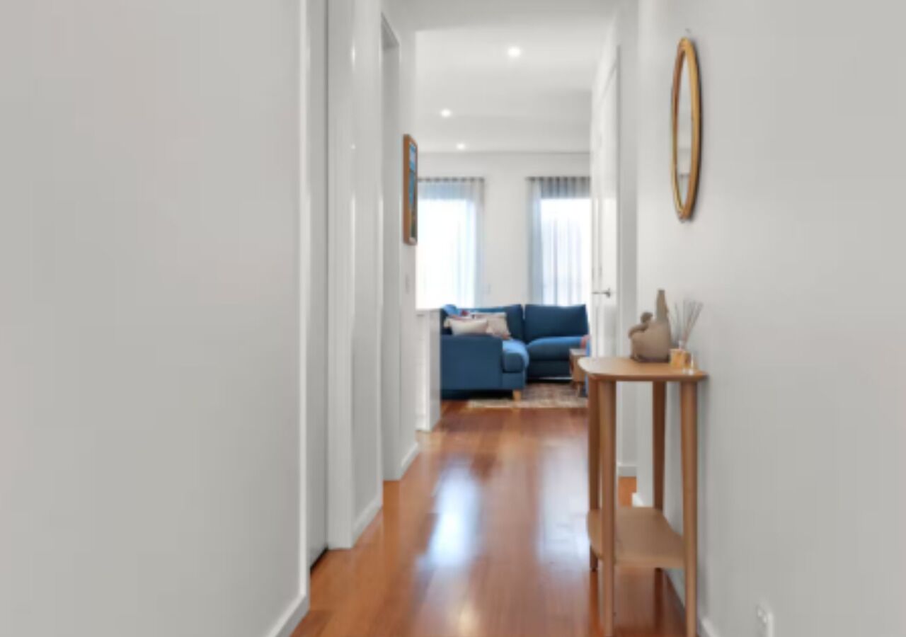 A hallway with a wooden floor leading to a living area with a blue sofa and a small table with a vase.