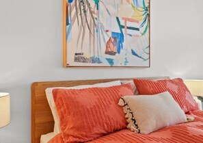 Un lit avec des oreillers rouges et beige, une tête de lit en bois et un tableau abstrait aux couleurs vives accroché au mur.