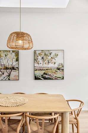 Ein Essbereich mit Holztisch, gewobenen Stühlen, einer gewobenen Pendelleuchte und zwei Landschaftsbildern an der Wand.