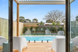 Ein Poolbereich mit Glasveranda, Holzterrasse und weißen Korbmöbeln.