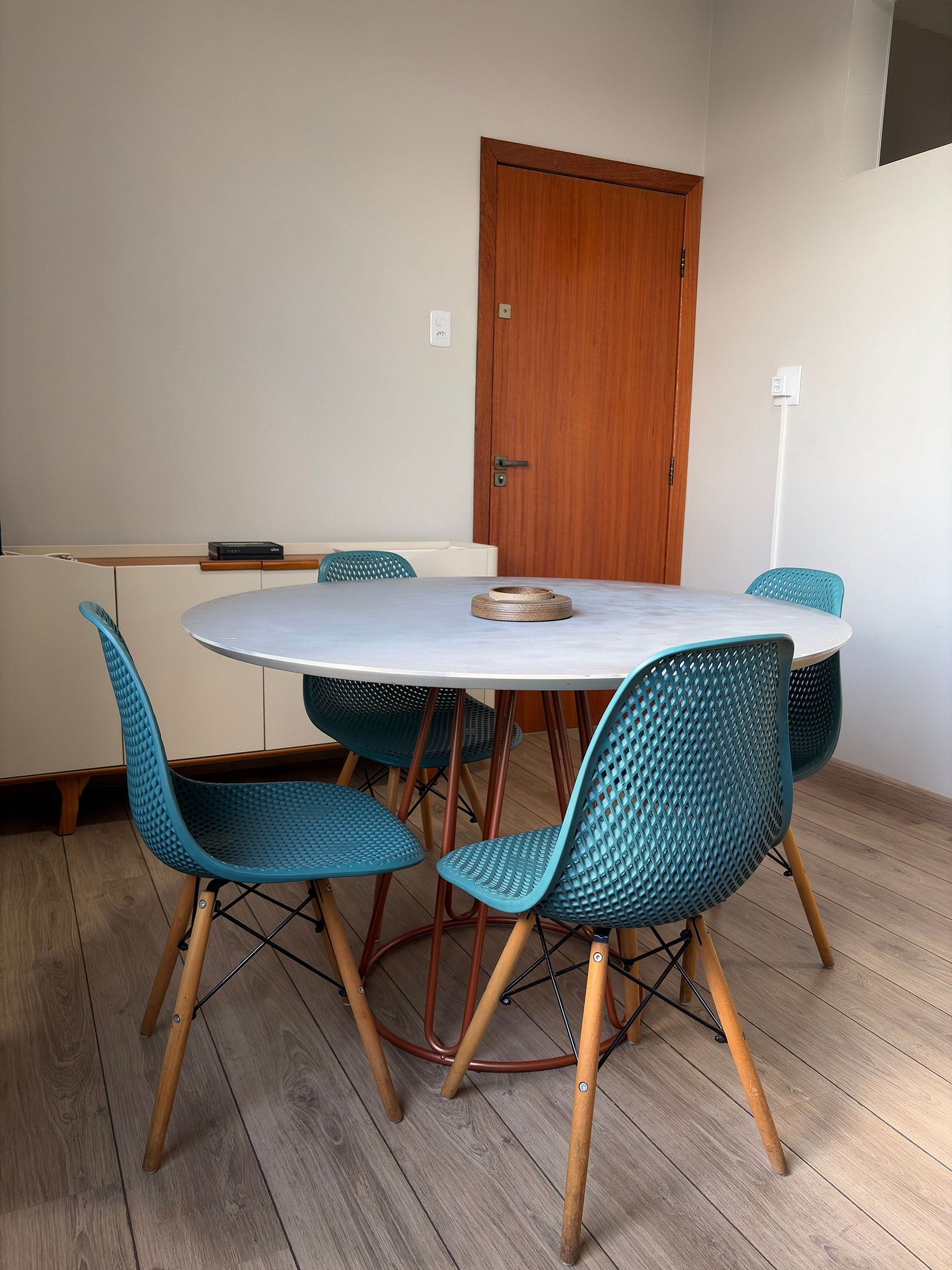 Sala de conferência com uma mesa redonda, quatro cadeiras, uma porta de madeira e uma parede branca.