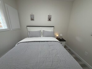 Een keurig opgemaakt bed met gestreepte lakens, een nachtkastje met een lamp en een klein spiegelbeeld, en twee aan de muur bevestigde fotolijstjes.