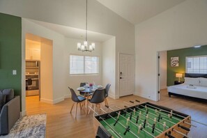 Een woonkamer met een tafelvoetbaltafel, een eetgedeelte met stoelen en een tafel, een keuken met apparatuur, en een slaapkamer met een bed en een nachtkastje.
