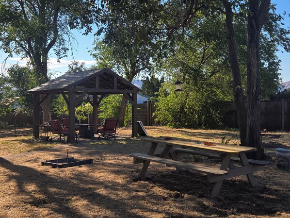 A wooden pavilion with a table and chairs, surrounded by trees and a picnic table.