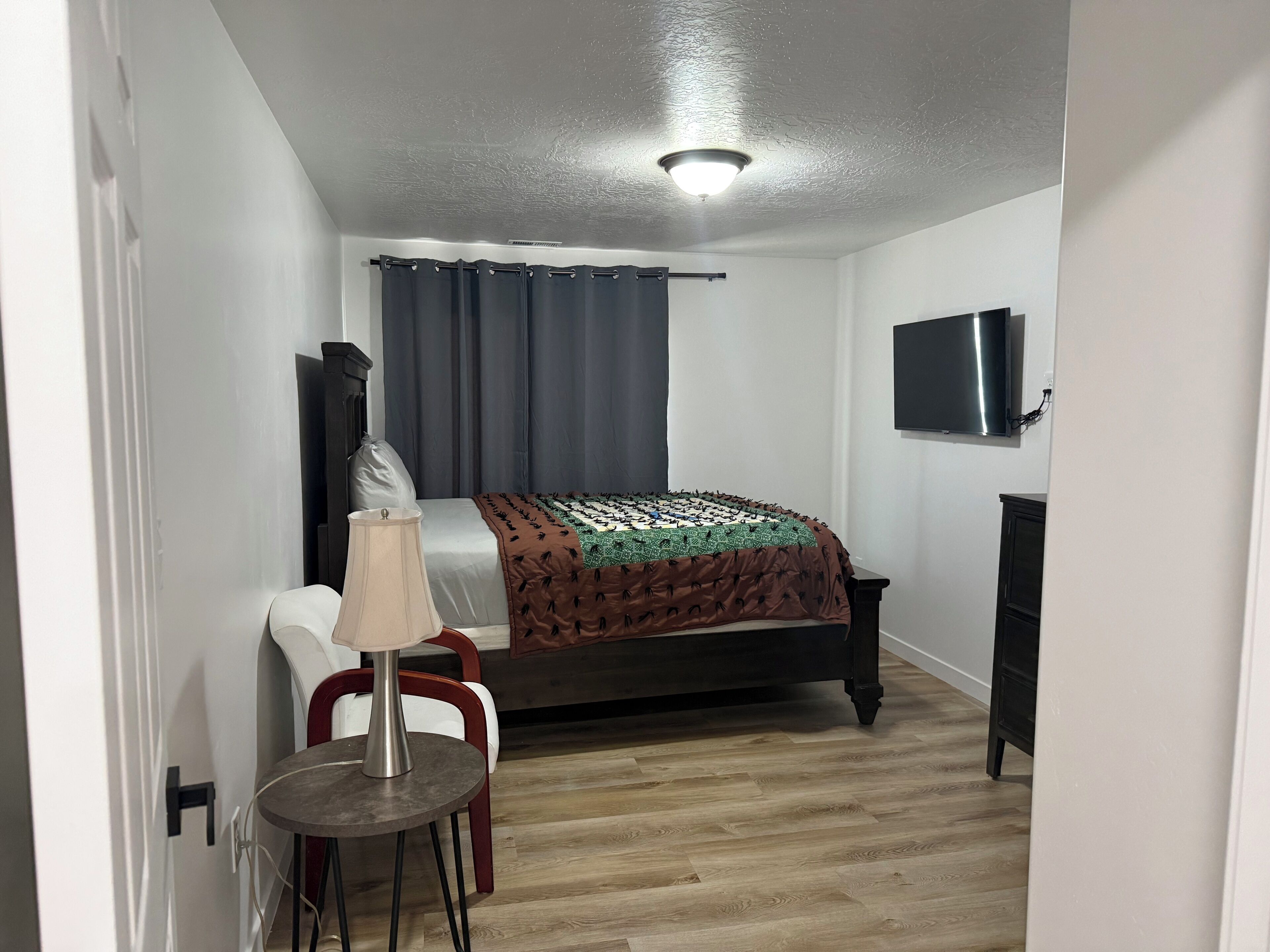 A bedroom with a bed, a TV mounted on the wall, a desk with a chair, and a small table with a lamp.