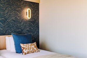 Una cama con ropa blanca, una almohada azul y otra almohada de diseño marrón y blanco, y una pared con un patrón ondulado.