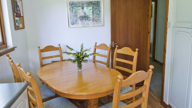 Een eetgedeelte met een houten tafel en stoelen, een vaas met bloemen, een ingelijst kunstwerk aan de muur en een deur naar een andere kamer.