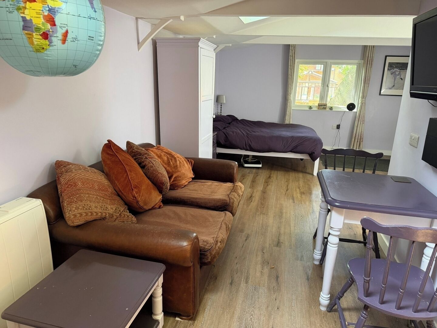 A compact living space with a brown leather sofa, a small table, a bed with a purple blanket, a desk with a chair, and a globe on the wall.