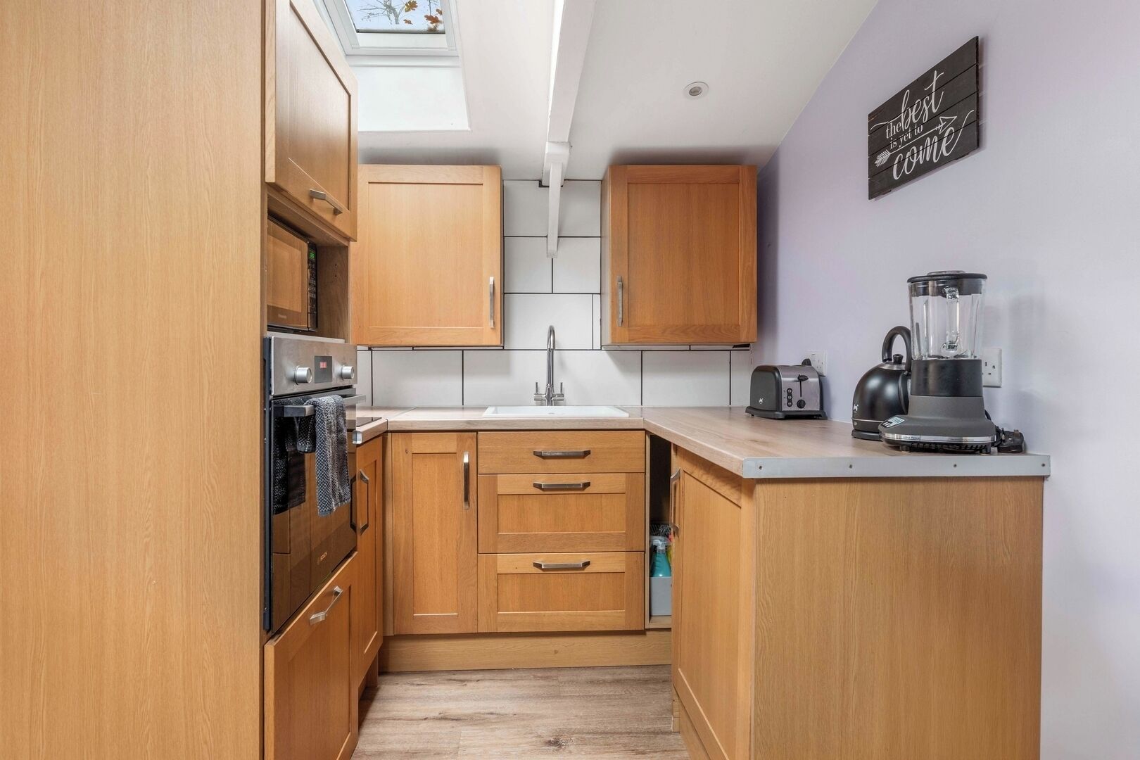 A compact kitchen with wooden cabinets, a built-in oven, a sink, a toaster, and a blender on the countertop.