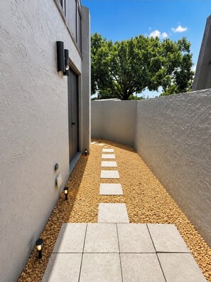 A narrow pathway with stepping stones leading to a building entrance, surrounded by pebbles and walls.