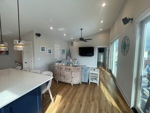 A modern kitchen with a large island, a dining area with a table and chairs, a built-in entertainment unit with a TV, and a large window providing natural light. - On the Intracoastal Waterway with a dock. Bring your boat, minutes to the beach (Oak Island)