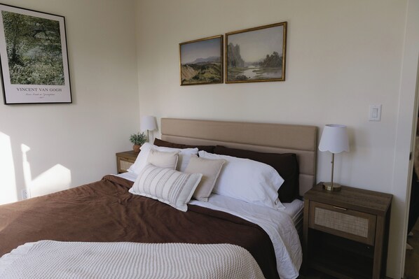 A neatly made bed with a brown headboard, two bedside tables with lamps, and two framed pictures on the wall. - The Vella | Private Modern Carriage House Near Shuswap Lake (Tappen)