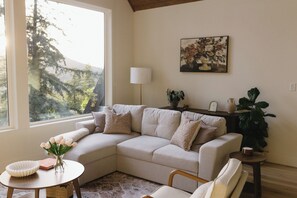 A living room with a sectional sofa, a round table, a vase with flowers, a lamp, and a painting on the wall. - The Vella | Private Modern Carriage House Near Shuswap Lake (Tappen)